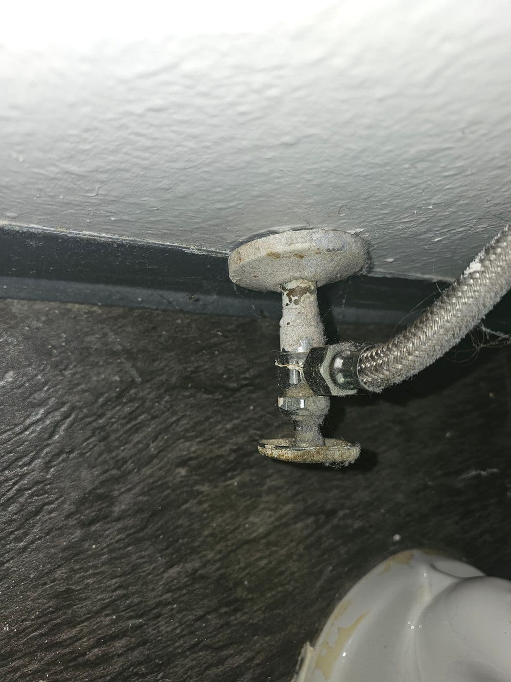 Close-up of a worn plumbing valve with a metal fitting and braided hose attachment.