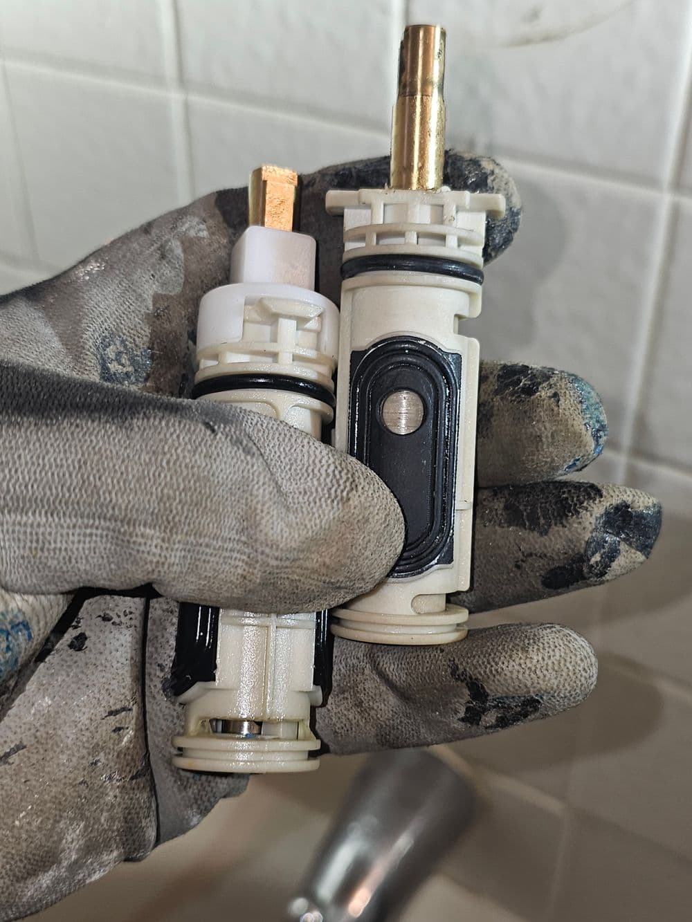 Person holding two plumbing cartridge valves with gloves in a bathroom setting.