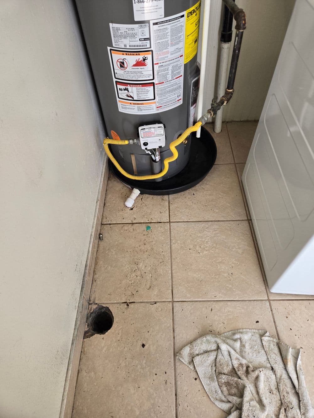 Water heater installation with drainage pipe and cleaning towel on tiled floor.
